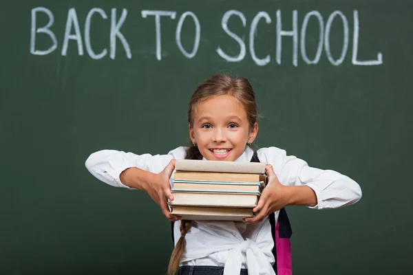 Aluna animado segurando pilha de livros perto de quadro-negro com volta à inscrição da escola — Fotografia de Stock