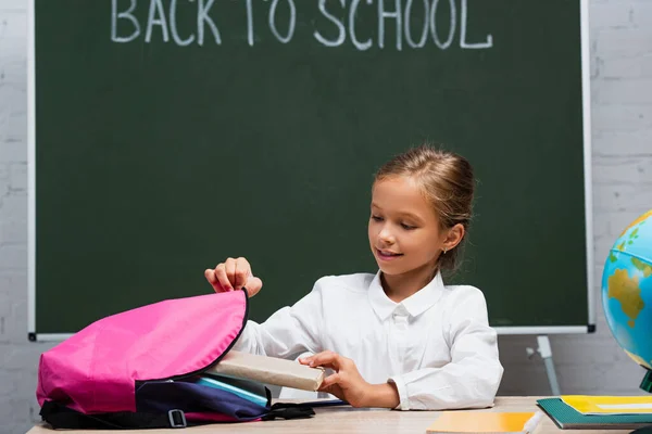 Estudante sorrindo levando livros de mochila enquanto sentado na mesa perto de quadro-negro com volta para a inscrição da escola — Fotografia de Stock