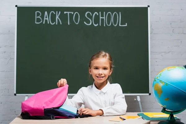 Estudante sorrindo levando livros de mochila enquanto sentado na mesa perto de quadro-negro com volta para a escola lettering — Fotografia de Stock