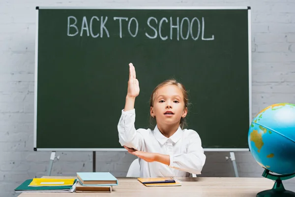 Entzückendes Schulmädchen hebt die Hand, während es am Schreibtisch in der Nähe von Globus und Kreidetafel mit dem Rücken zur Schule Inschrift sitzt — Stockfoto