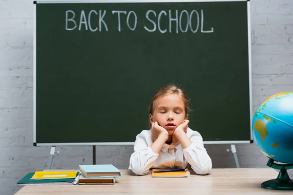 Scolaretta stanca seduta con gli occhi chiusi alla scrivania vicino al globo e alla lavagna con iscrizione di ritorno a scuola — Foto stock