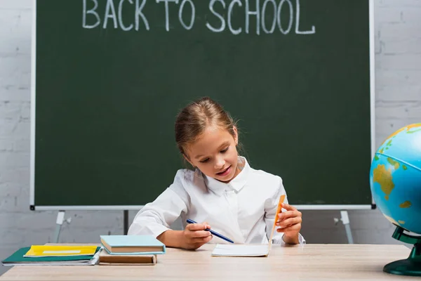 Attentive schoolgirl looking in copy book while sitting at desk near textbooks and chalkboard with back to school inscription — Stock Photo