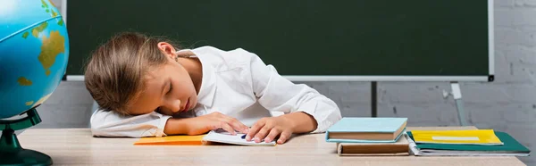 Imagem horizontal de estudante cansada dormindo na mesa perto do globo e quadro-negro com as costas para a inscrição da escola — Fotografia de Stock