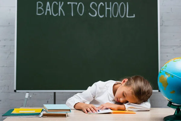 Erschöpfte Schülerin schläft am Schreibtisch neben Erdkugel und Tafel mit Schulbuchstaben — Stockfoto