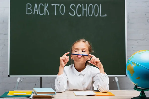 Linda colegiala sosteniendo pluma cerca de los labios mientras está sentado en el escritorio cerca de globo y pizarra con letras de vuelta a la escuela - foto de stock