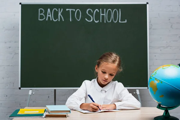 Concentrated schoolgirl writing in copy book while sitting at desk near globe and chalkboard with back to school lettering — Stock Photo