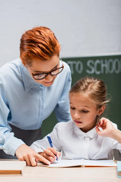 Внимательный учитель указывает пальцем на симпатичную школьницу, пишущую в книжке — стоковое фото