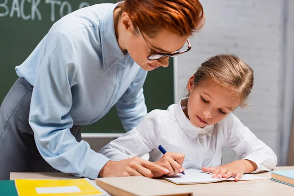 Attentive teacher pointing with finger near schoolgirl writing in notebook — Stock Photo