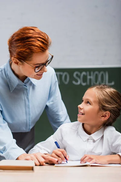 Glückliches Schulmädchen schaut attraktive Lehrerin beim Schreiben in Kopierbuch an — Stockfoto