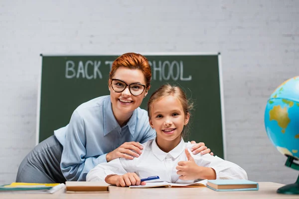 Fuoco selettivo di insegnante sorridente che tocca spalle di scolaretta felice che scrive in notebook — Foto stock