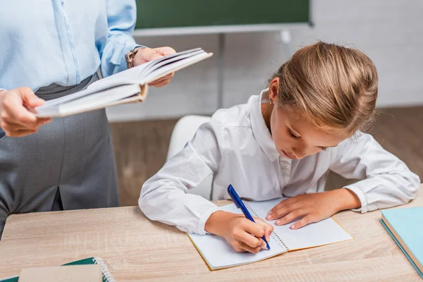 Partial view of teacher with book near schoolgirl sitting at desk and writing in copy book — Stock Photo