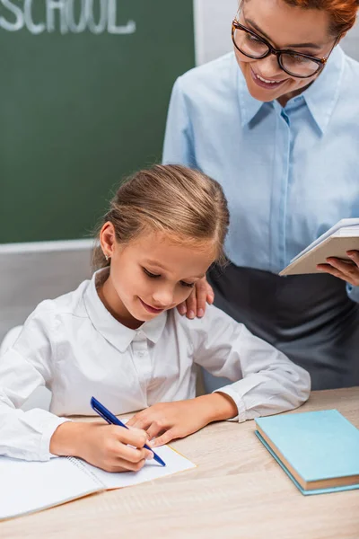 Enseignant souriant regardant écolière assise au bureau et écrivant dans un cahier — Photo de stock