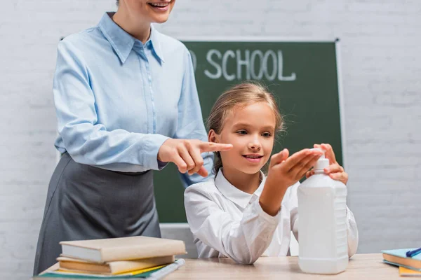 Partial view of teacher pointing with finger near schoolgirl applying sanitizer on hands — Stock Photo