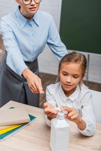 Vue partielle du professeur pointant avec le doigt près de l'écolière appliquant un désinfectant pour les mains — Photo de stock