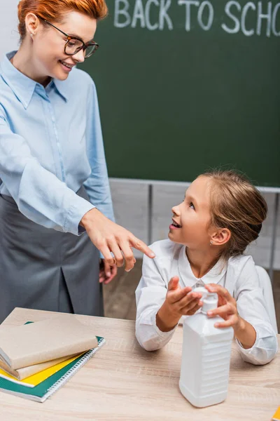 Enfoque selectivo de profesor sonriente señalando con el dedo cerca de colegiala aplicación de mano antiséptico - foto de stock