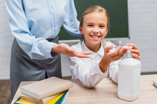 Vue recadrée du professeur près de l'écolière souriante appliquant un désinfectant sur les mains — Photo de stock