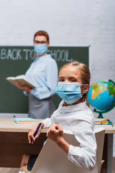 Foyer sélectif de l'écolière en masque médical regardant la caméra, et enseignant debout près de tableau noir avec livre — Photo de stock