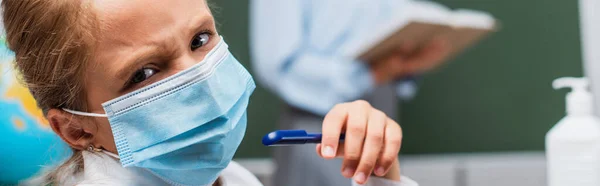 Selective focus of displeased schoolgirl in medical mask looking at camera, while teacher standing on background, panoramic crop — Stock Photo