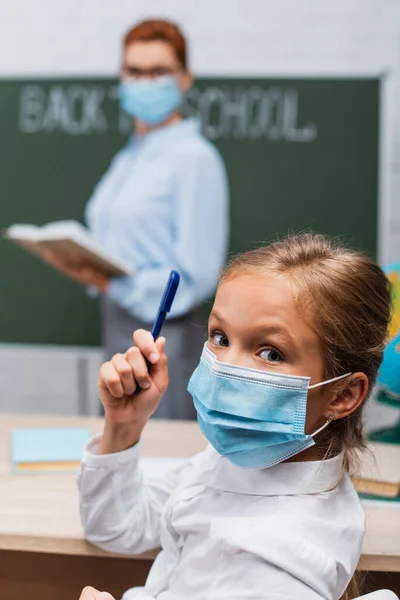 Foyer sélectif de l'écolière dans le masque médical tenant stylo et regardant la caméra, et enseignant avec livre près du tableau — Photo de stock