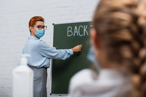 Vue arrière de l'écolière, et professeur dans l'écriture de masque médical sur tableau noir — Photo de stock