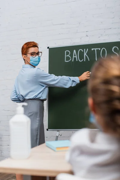 Vista posteriore di scolara, e insegnante in maschera medica scrittura sulla lavagna — Foto stock