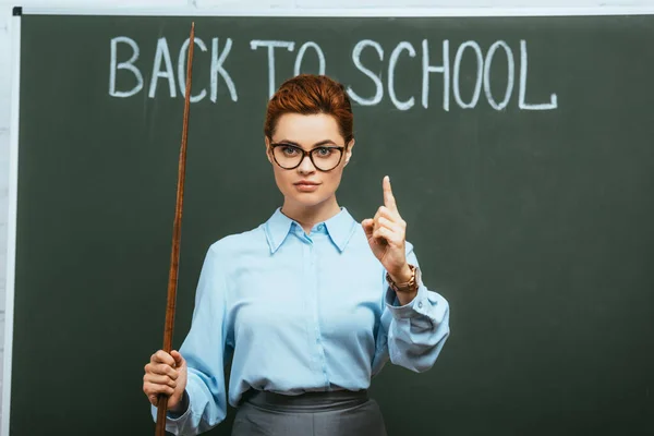 Ernsthafter Lehrer hält Zeigestab in der Hand und zeigt Aufmerksamkeitsgeste in der Nähe der Tafel mit Schulaufschrift — Stockfoto