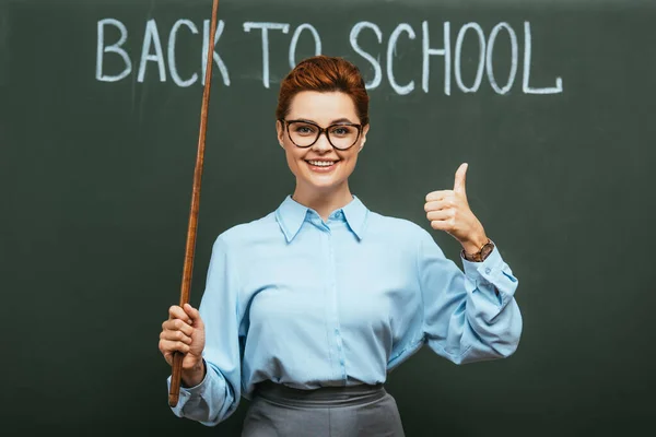 Smiling teacher with pointing stick showing thumb up near chalkboard with back to school lettering — Stock Photo