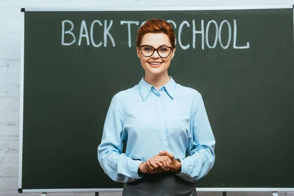 Cheerful teacher standing with clenched hands near chalkboard with back to school inscription — Stock Photo