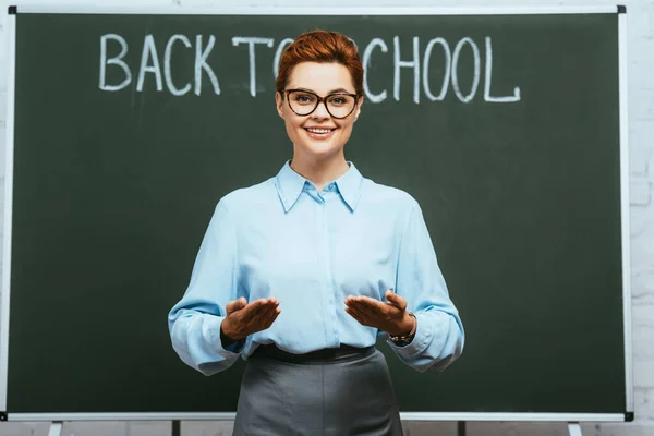 Professor feliz de pé com os braços abertos perto de quadro-negro com volta para a escola lettering — Fotografia de Stock