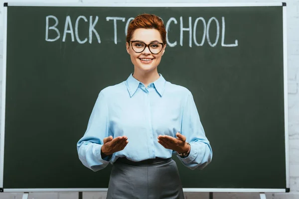 Profesor feliz de pie con los brazos abiertos cerca de pizarra con letras de vuelta a la escuela - foto de stock