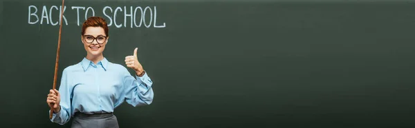 Concepto panorámico de profesor sonriente con palo de apuntar mostrando el pulgar hacia arriba cerca de pizarra con letras de vuelta a la escuela - foto de stock