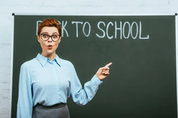 Surprised teacher pointing with finger near chalkboard with back to school inscription — Stock Photo
