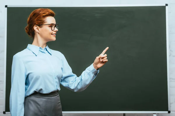 Smiling teacher looking away and pointing with finger near chalkboard — Stock Photo