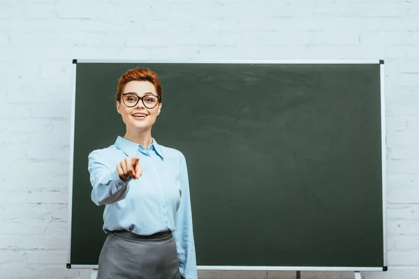 Smiling teacher looking at camera and pointing with finger near chalkboard — Stock Photo