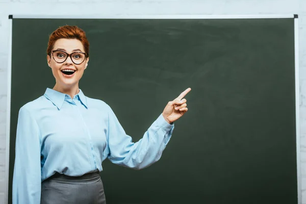 Excited teacher looking at camera and pointing with finger near chalkboard — Stock Photo