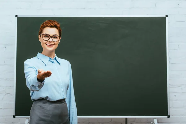 Happy teacher smiling at camera while standing with open arm near chalkboard — Stock Photo