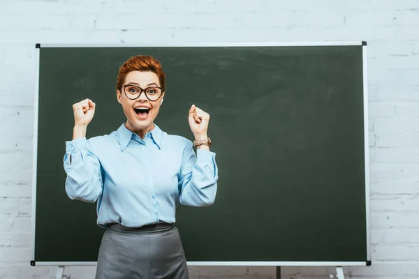 Profesor emocionado mostrando gesto ganador y mirando a la cámara mientras está de pie cerca de pizarra - foto de stock