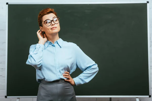 Enseignant réfléchi touchant la tête et regardant loin tout en se tenant avec la main sur la hanche près du tableau — Photo de stock