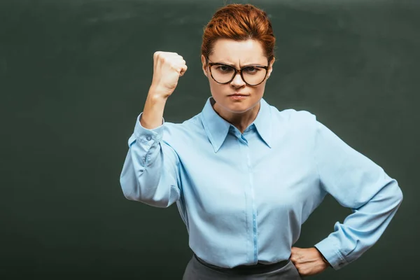 Professeur irrité montrant poing serré tout en se tenant avec la main sur la hanche près du tableau — Photo de stock