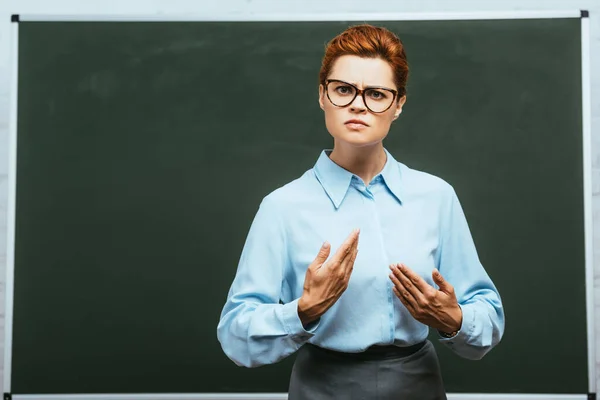 Seriöser Lehrer mit Brille blickt in die Kamera, während er neben der Tafel steht — Stockfoto