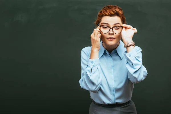 Young teacher touching eyeglasses and eye while standing near chalkboard — Stock Photo