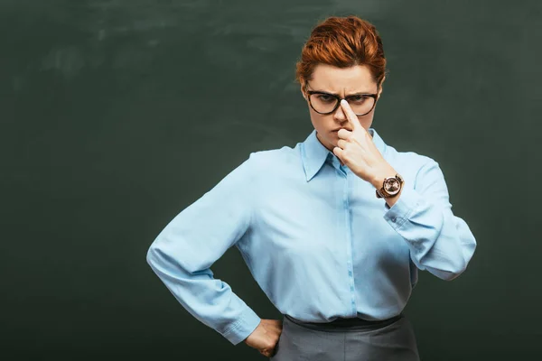 Enseignant sérieux touchant des lunettes tout en se tenant avec la main sur la hanche près du tableau noir — Photo de stock