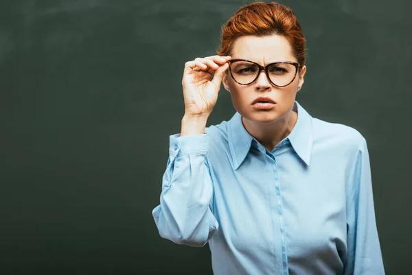 Concentrated teacher touching eyeglasses while peering near chalkboard — Stock Photo