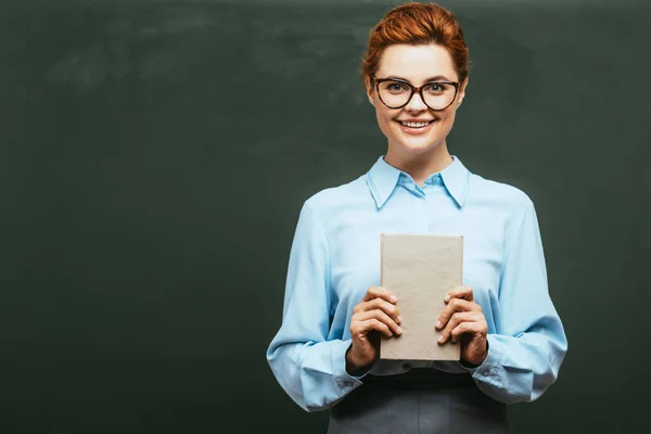 Happy teacher in eyeglasses holding book while standing near chalkboard — Stock Photo