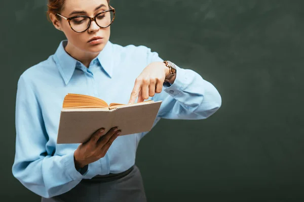 Aufmerksame Lehrerin in Brille zeigt mit dem Finger auf aufgeschlagenes Buch, während sie in der Nähe der Tafel steht — Stockfoto