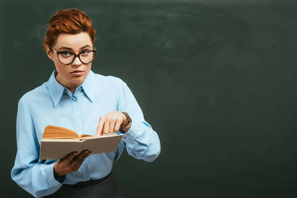 Aufmerksame Lehrerin mit Brille zeigt auf aufgeschlagenes Buch, während sie neben Kreidetafel steht — Stockfoto
