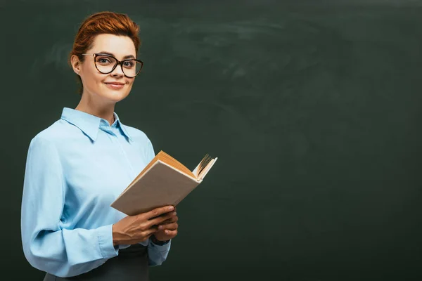 Happy, beautiful teacher in eyeglasses holding open book while standing near chalkboard — Stock Photo