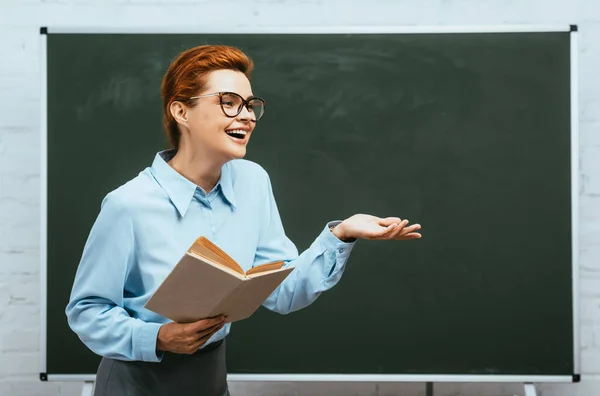 Laughing teacher in eyeglasses holding book while standing near chalkboard with open arm — Stock Photo