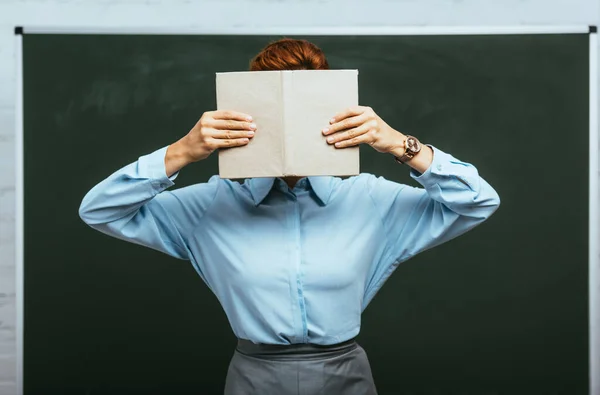 Profesor en blusa azul oscureciendo la cara con libro mientras está de pie cerca de pizarra - foto de stock