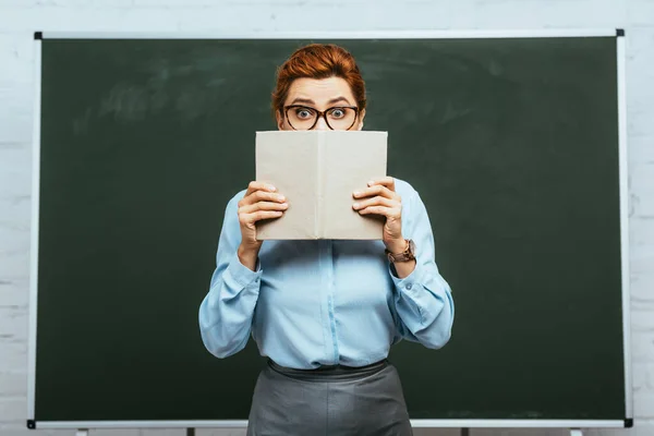 Scared teacher in eyeglasses covering face with open book while standing near chalkboard — Stock Photo